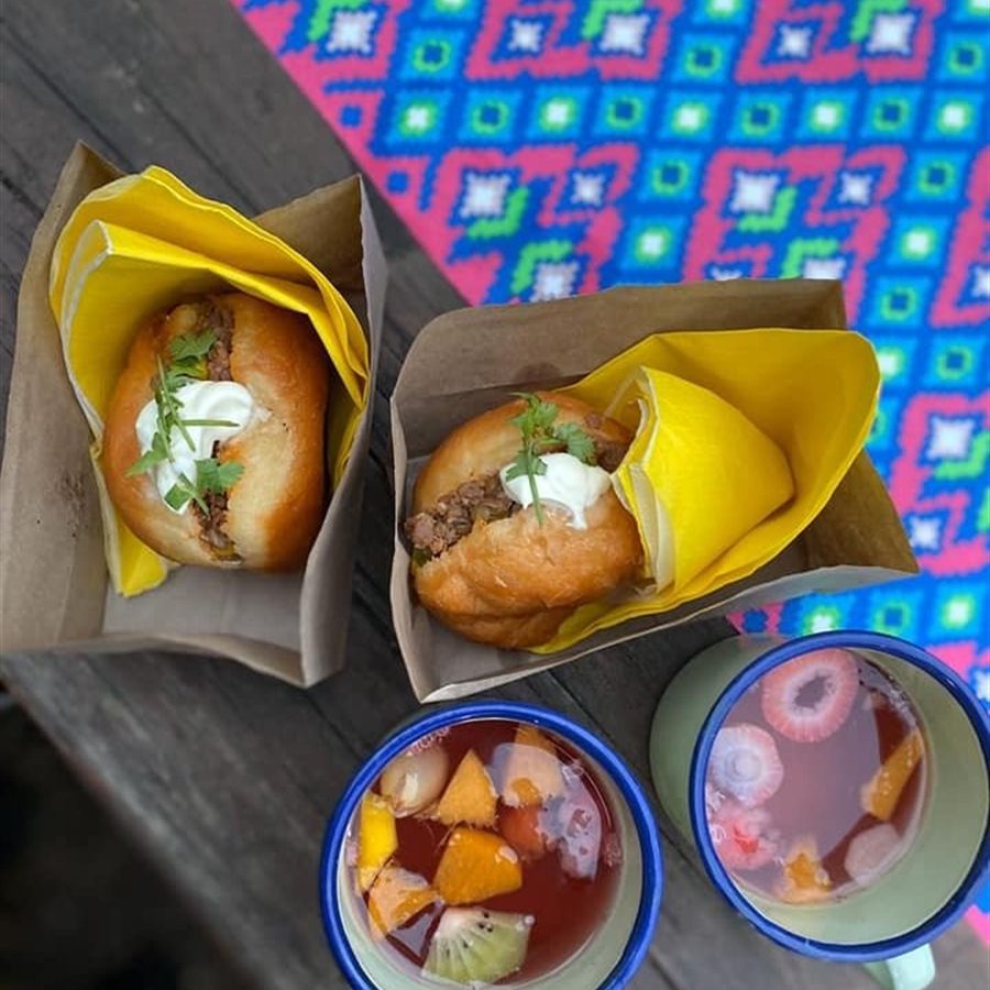 Two filled rolls in paper wrapping beside enamel mugs of iced fruit punch on a wooden table.