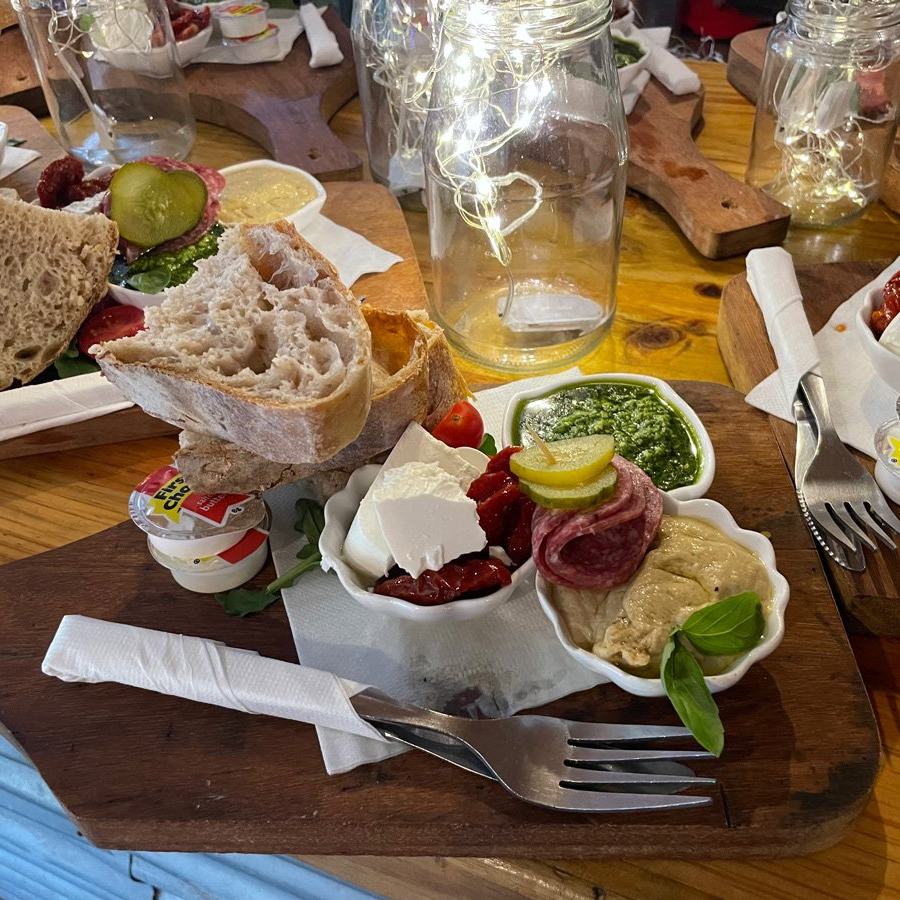 A wooden board with fresh bread, cheese, cured meats, pesto and hummus, lit by fairy lights in glass jars.