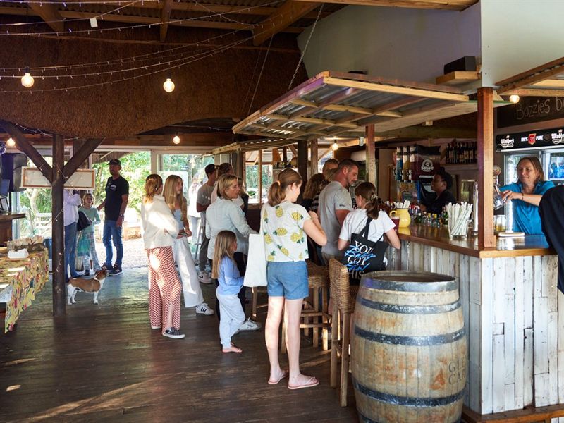 Guests queuing at the wooden bar under a thatched roof, with a wine barrel and strings of fairy lights overhead.